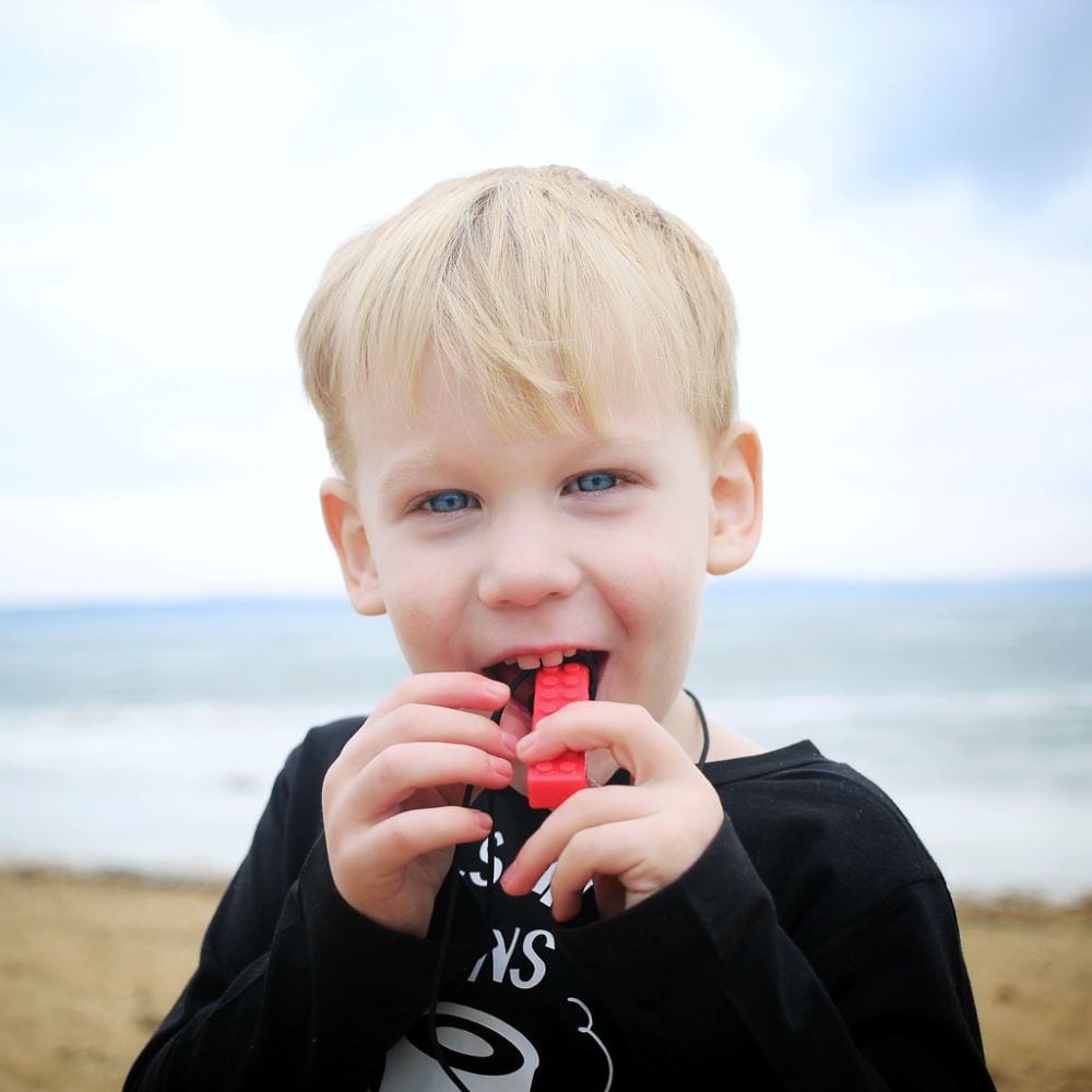 Caucasian toddler chewing on Ark's brick stick textured sensory chew necklace in red