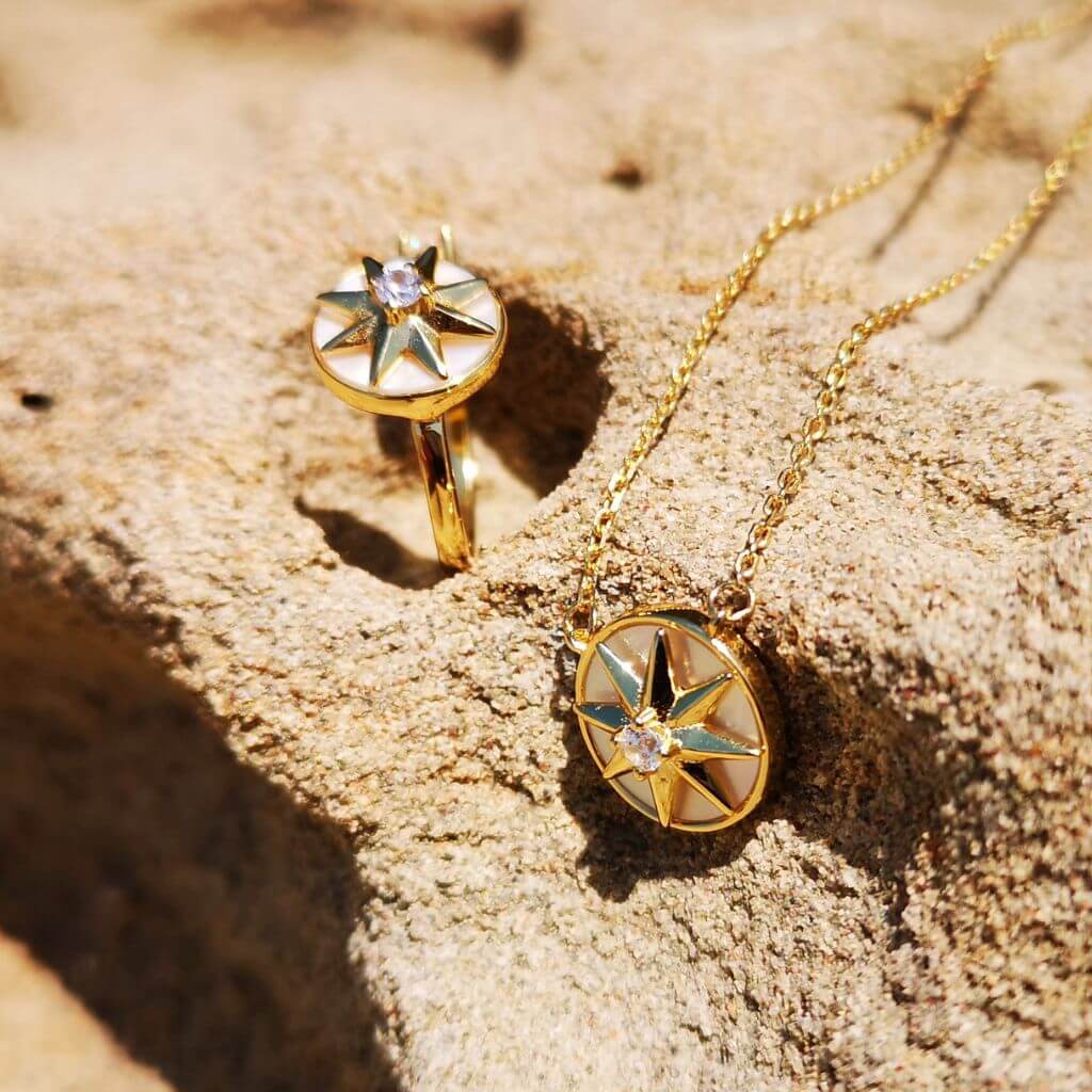 Gold-tone jewelry set featuring a compass rose pendant necklace and matching stud earring, both with a central clear gemstone, displayed on a textured sandy surface.
