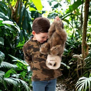 Caucasian girl holding a baby sloth hand puppet from Folkmanis from its legs on forest background