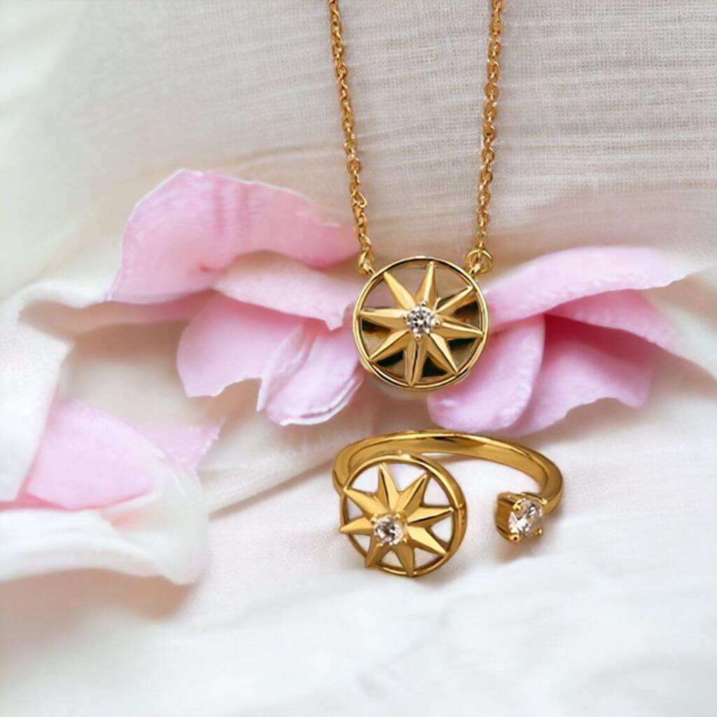 Gold-toned jewelry set featuring a necklace and matching open ring, both with starburst-shaped pendants and small clear stones, styled on soft white fabric with pink petals.