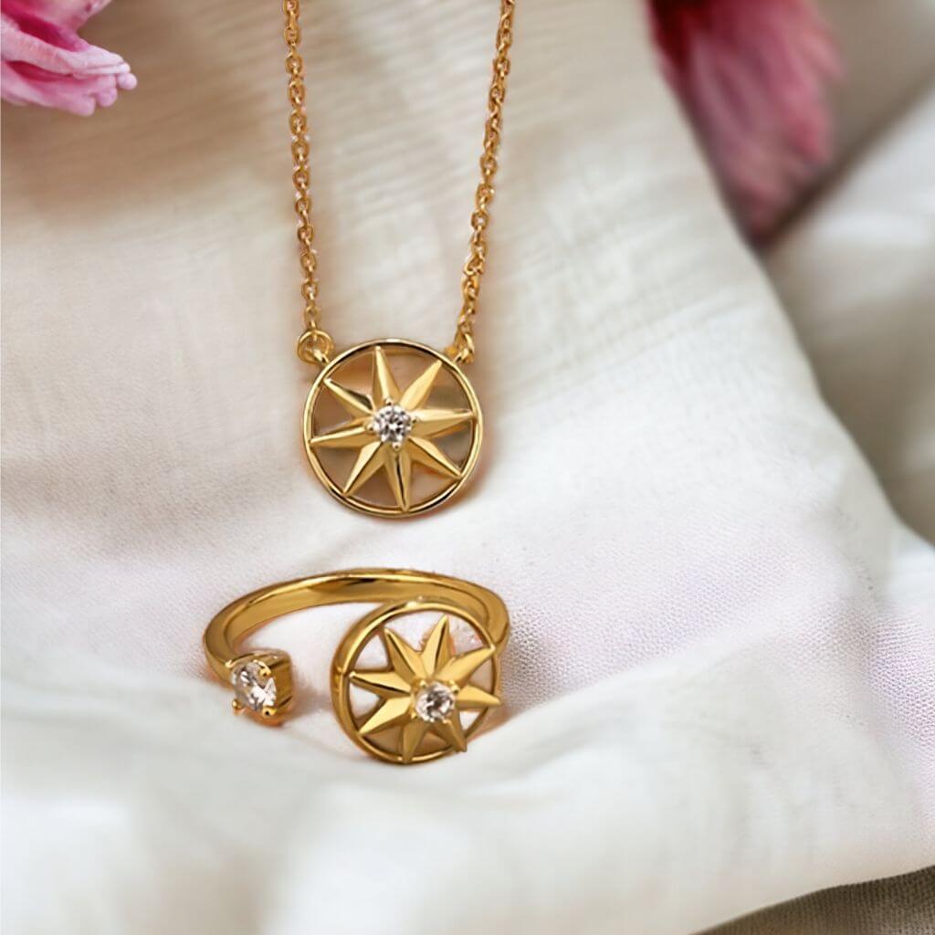 Gold jewelry set featuring a necklace and matching ring, both with circular starburst pendants and small clear gemstones, displayed on soft white fabric.