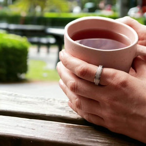 Woman's hands holding a tea mug wearing a silver fidget ring on the index finger on green blurred background