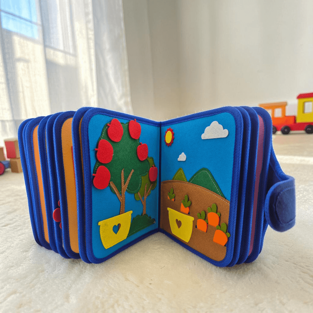 Felt quiet book open on white rug showing apple tree page and vegetable garden with carrots, wooden toy train in background