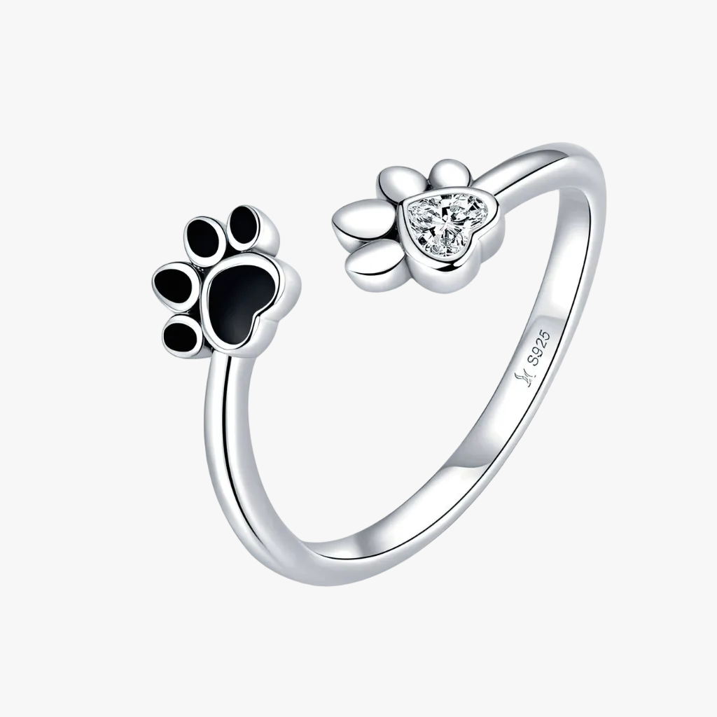 A sterling silver open ring designed for pets, featuring one paw print with black enamel and the other with a clear heart-shaped crystal, on a light gray background.