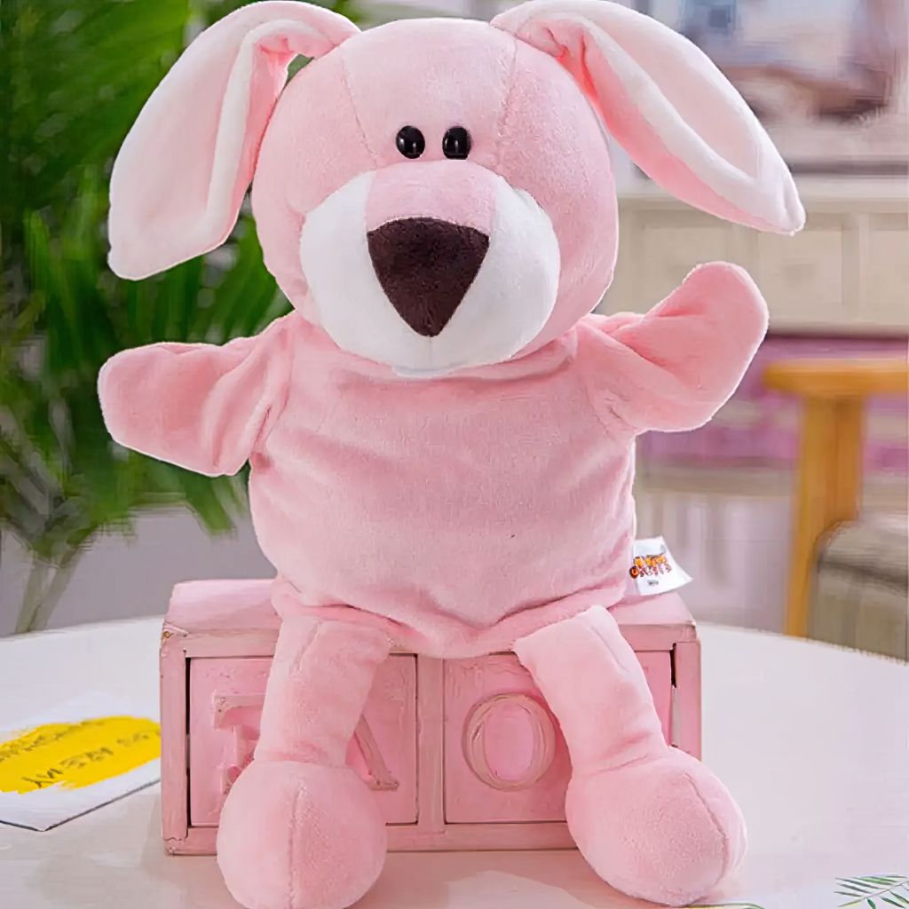 Pink bunny hand puppet seated on a decorative pink wooden box on a white table, showing full body with long floppy ears, white muzzle and dark nose, in a bright lifestyle setting