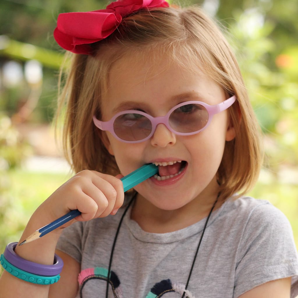Toddler girl with glasses and ribbon in her hair chewing on  a teal Ark&#39;s krypto bite chew pencil topper