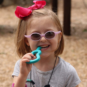Caucasian toddler girl wearing glasses chewing on Ark's guitar shaped textured sensory chew