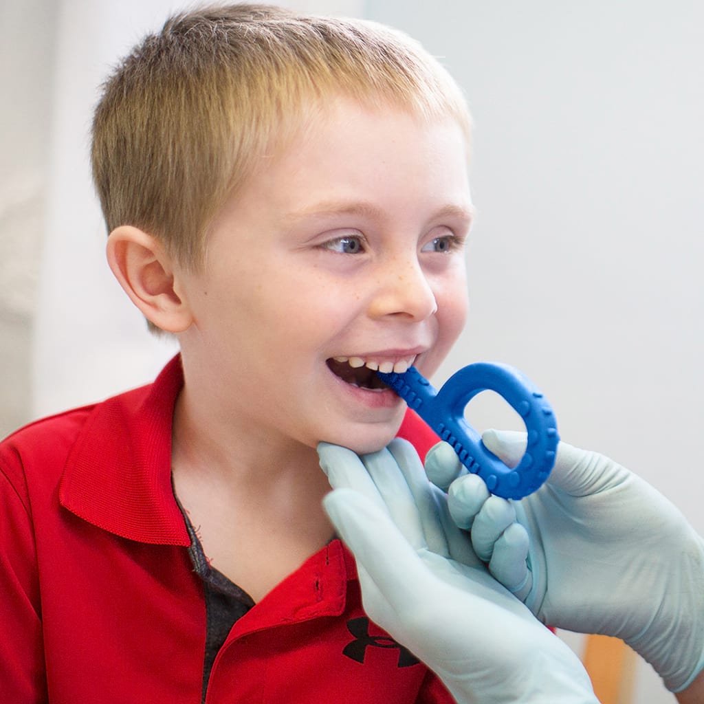 Caucasian boy chewing on Ark's textured grabber held by a speech language pathologist during chewing therapy